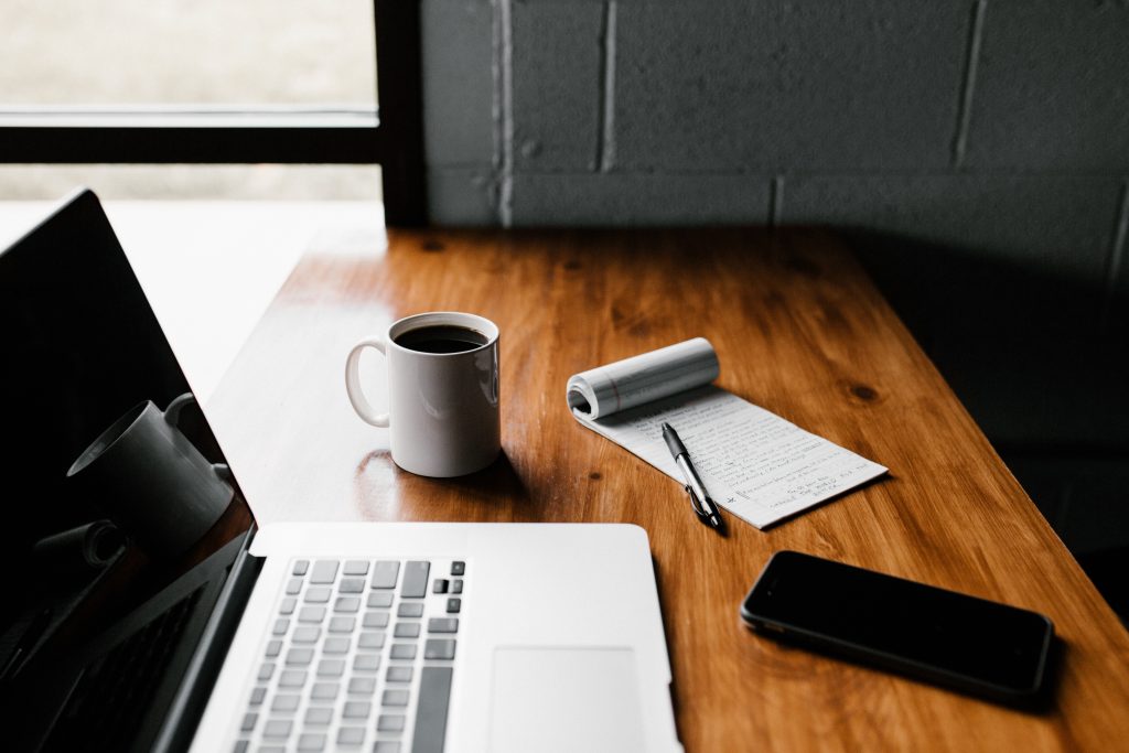 Laptop on desk with notepad, pen, mobile device and mug.