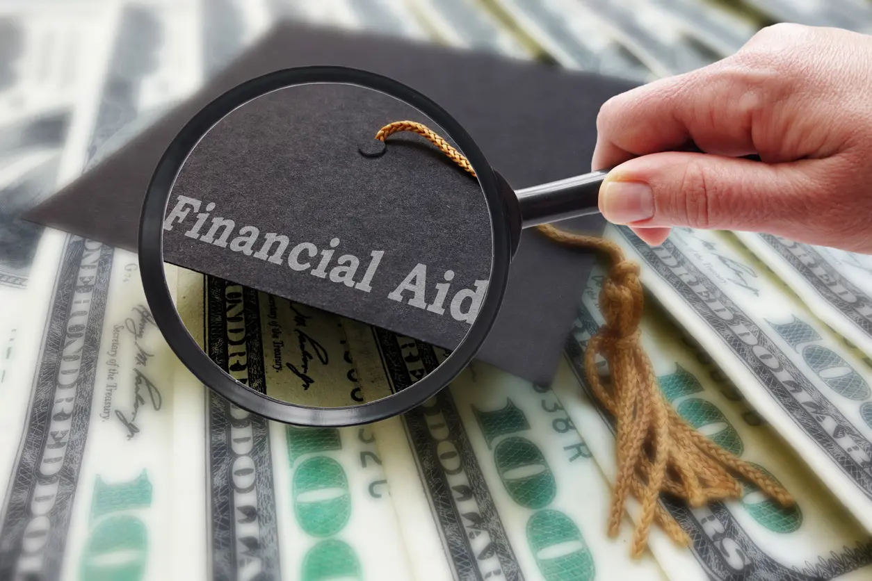 Magnifying glass highlighting “Financial Aid” text over U.S. dollar bills and a graduation cap
