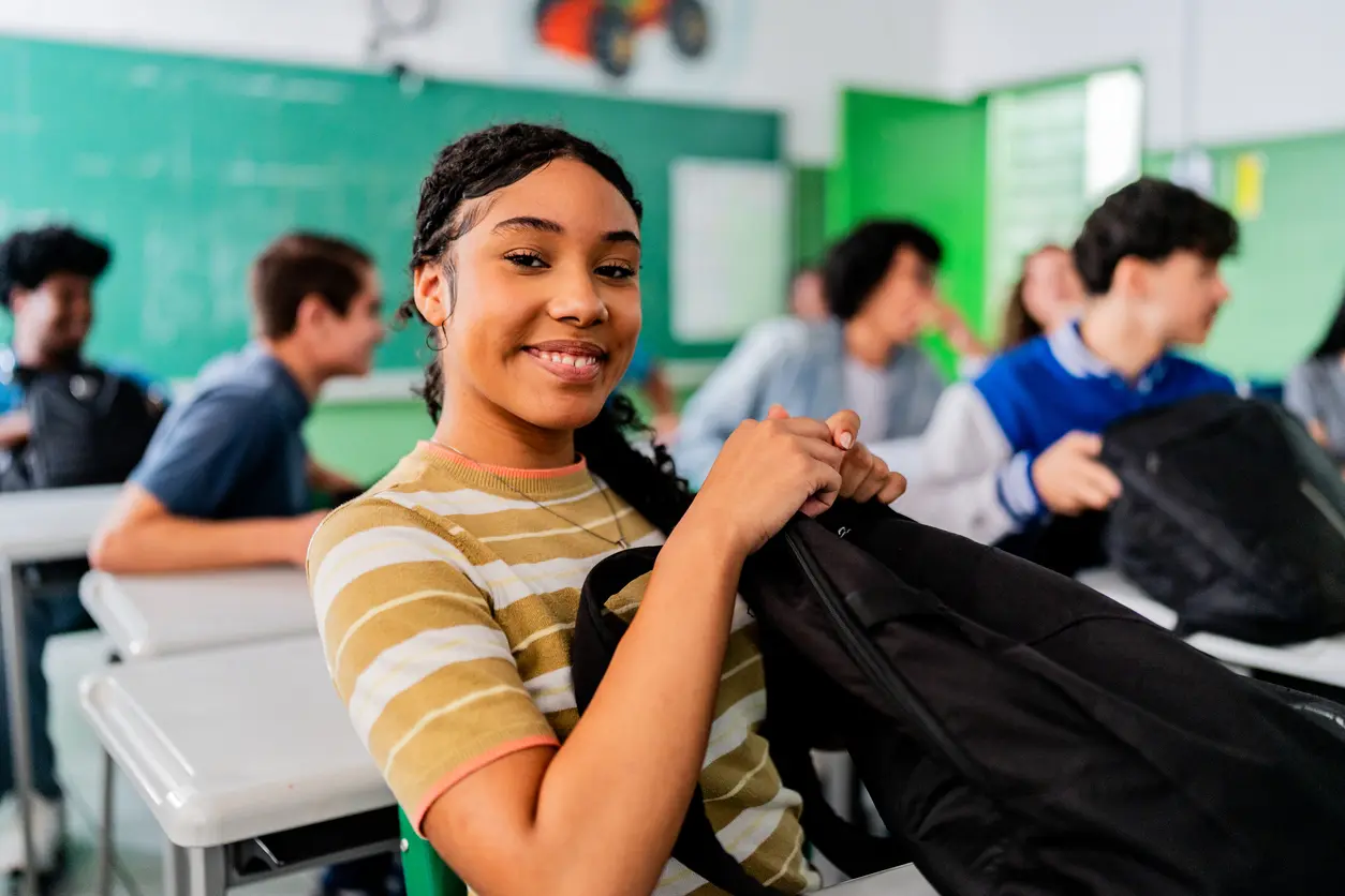 High school student smiling at desk while organizing backpack in classroom